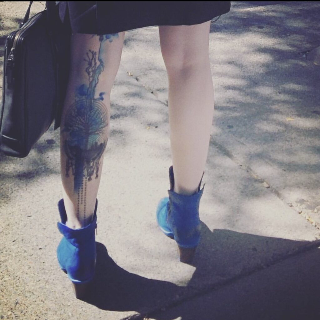 The backs of some very pale legs with blue shoes, and a watercolor tattoo with a tree, a maze, and skeletons on it on the back of the left leg from ankle to disappearing under a dress.