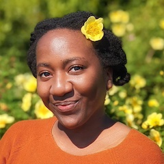 A smiling woman with dark hair with a yellow flower in it wearing an orange top outdoors in a sunny pastoral setting.