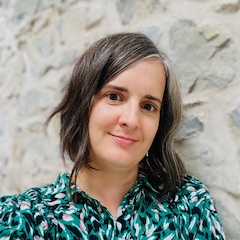 A smiling light-skinned woman with medium length hair and a colourful green top in front of a stucco wall.