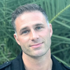 A short-haired white man with a chiselled jaw tilts his head to one side and looks dreamily out from in front of green foliage.