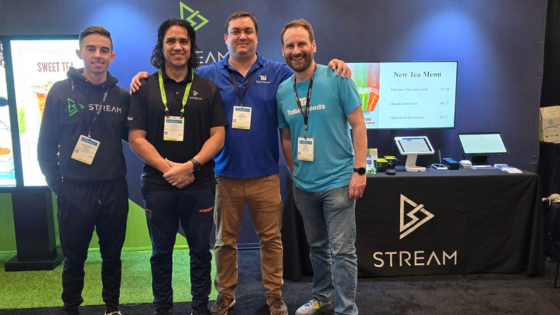 Photo of 4 men standing close together and smiling at an industry tradeshow. Behind them are displays for Table Needs and Stream