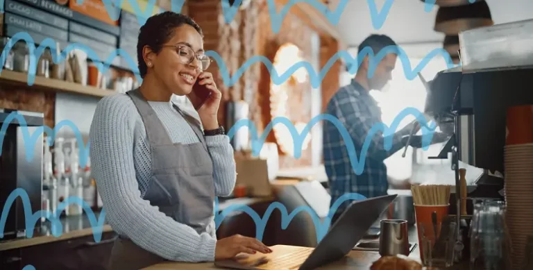 A smiling female bagel shop owner talks on the phone while working on a laptop in her shop, illustrating the importance of effective management practices for restaurant profitability.