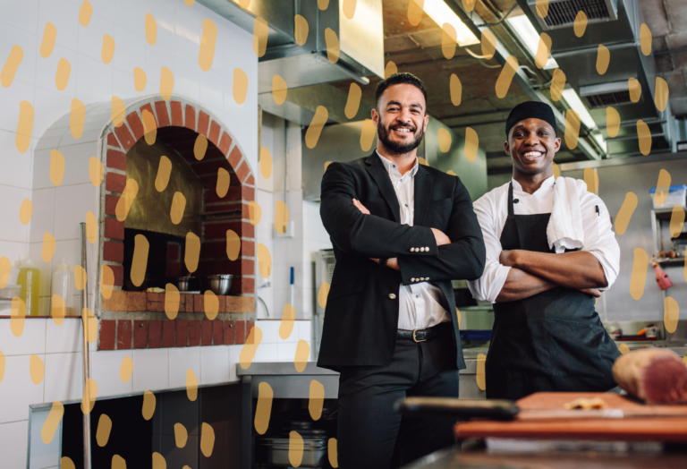 Two smiling men with their arms crossed standing in front of a brick oven in a restaurant