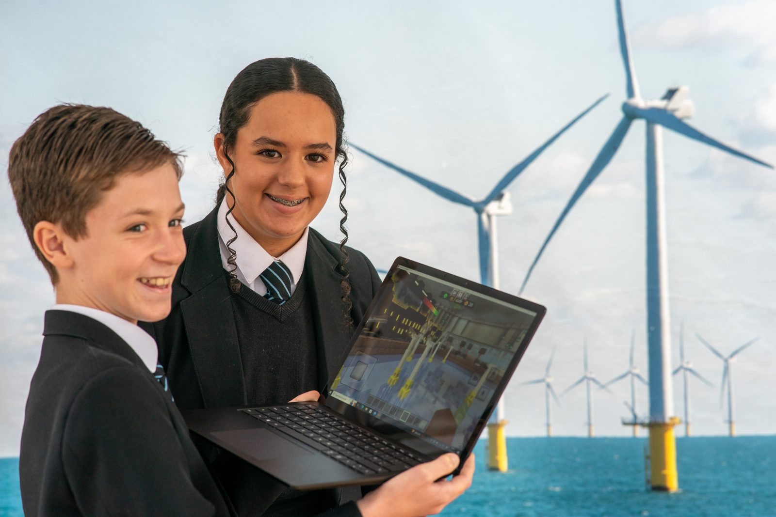 Two 12 year old students from Peacehaven Community School hold a laptop which shows a game of Minecraft on the screen portraying a wind farm. Behind them is a poster with a photograph of Rampion Wind Farm.