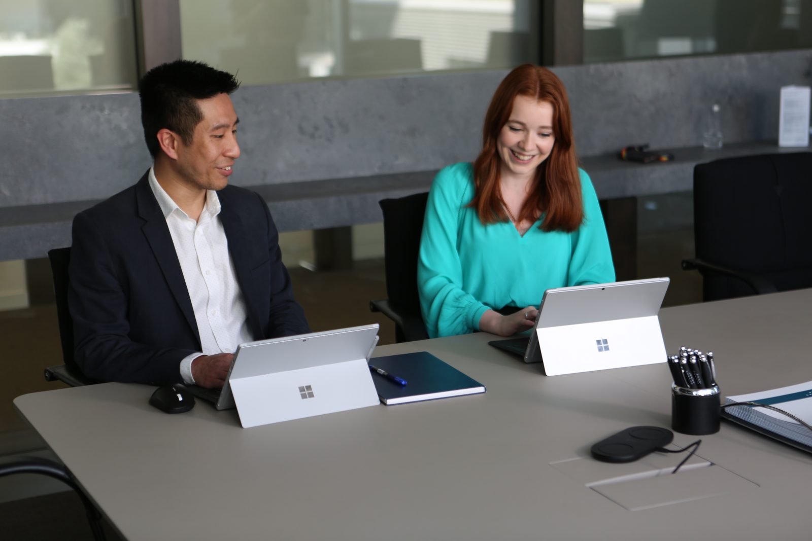 A man and a woman in an office sit at a desk with a Surface device each. They are smiling as they do work on their Surface devices.