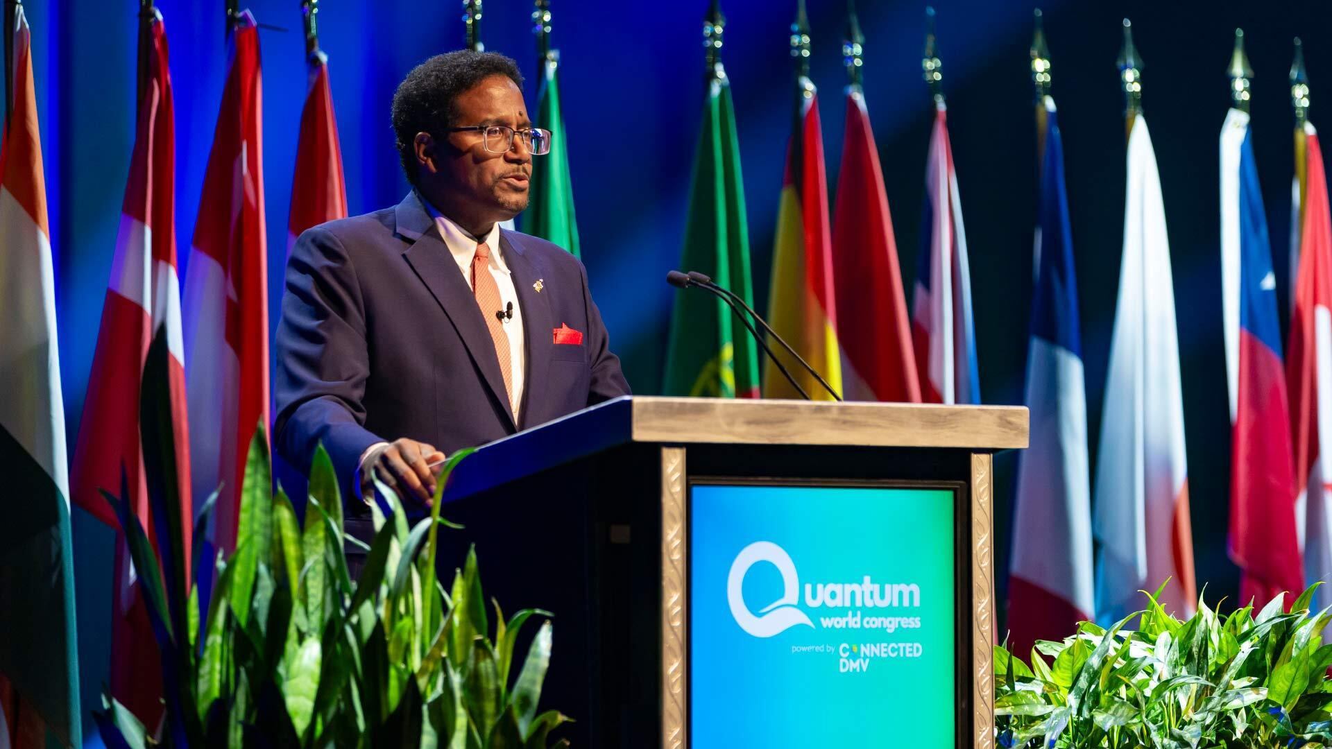 man at podium speaks in front of backdrop mentioning quantum