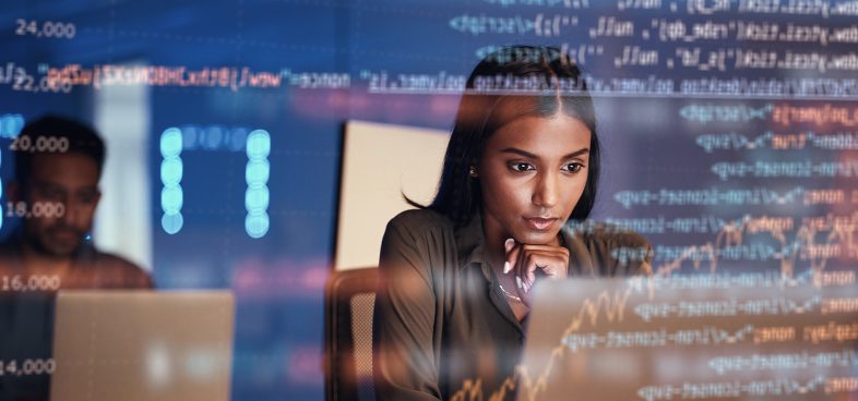 A woman is intently working on a computer in a modern office environment, surrounded by screens displaying dynamic digital data and stock market numbers, highlighting a focus on financial analysis.