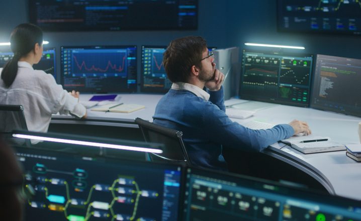 Representation of accelerating malware analysis. Two professionals working in a high-tech monitoring room filled with large screen displays showing data and graphs. One person is seated looking at a monitor, while the other, wearing glasses, is deep in thought.