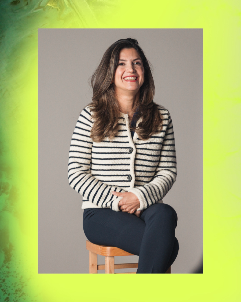 A woman with long brown hair wearing a striped sweater and dark pants, sitting on a wooden stool and smiling.