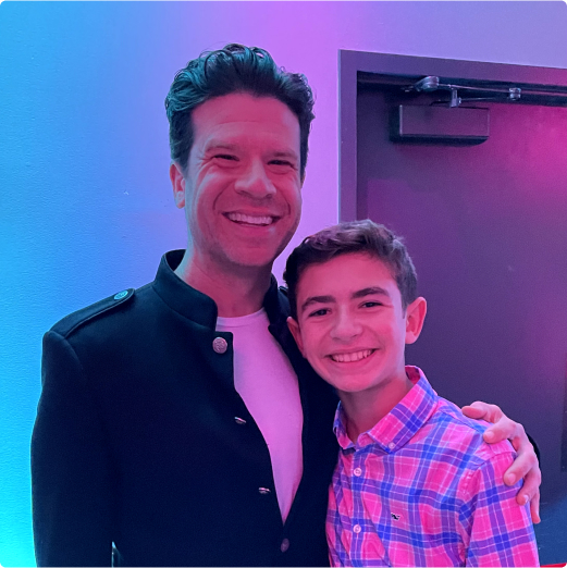 Two smiling males, an adult and a teenager, posing for a photo in a room with purple lighting. the adult wears a dark jacket, and the teen is in a plaid shirt.