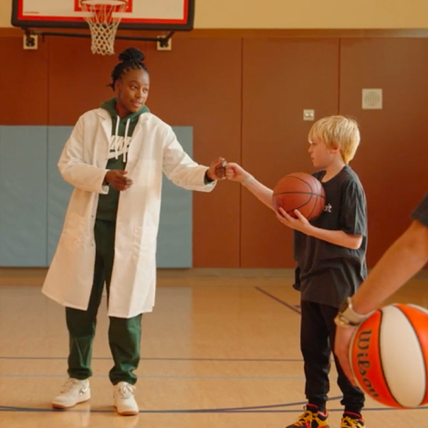 Two individuals wearing lab coats in a gym, one handing a basketball to the other, as they engage in a friendly interaction.