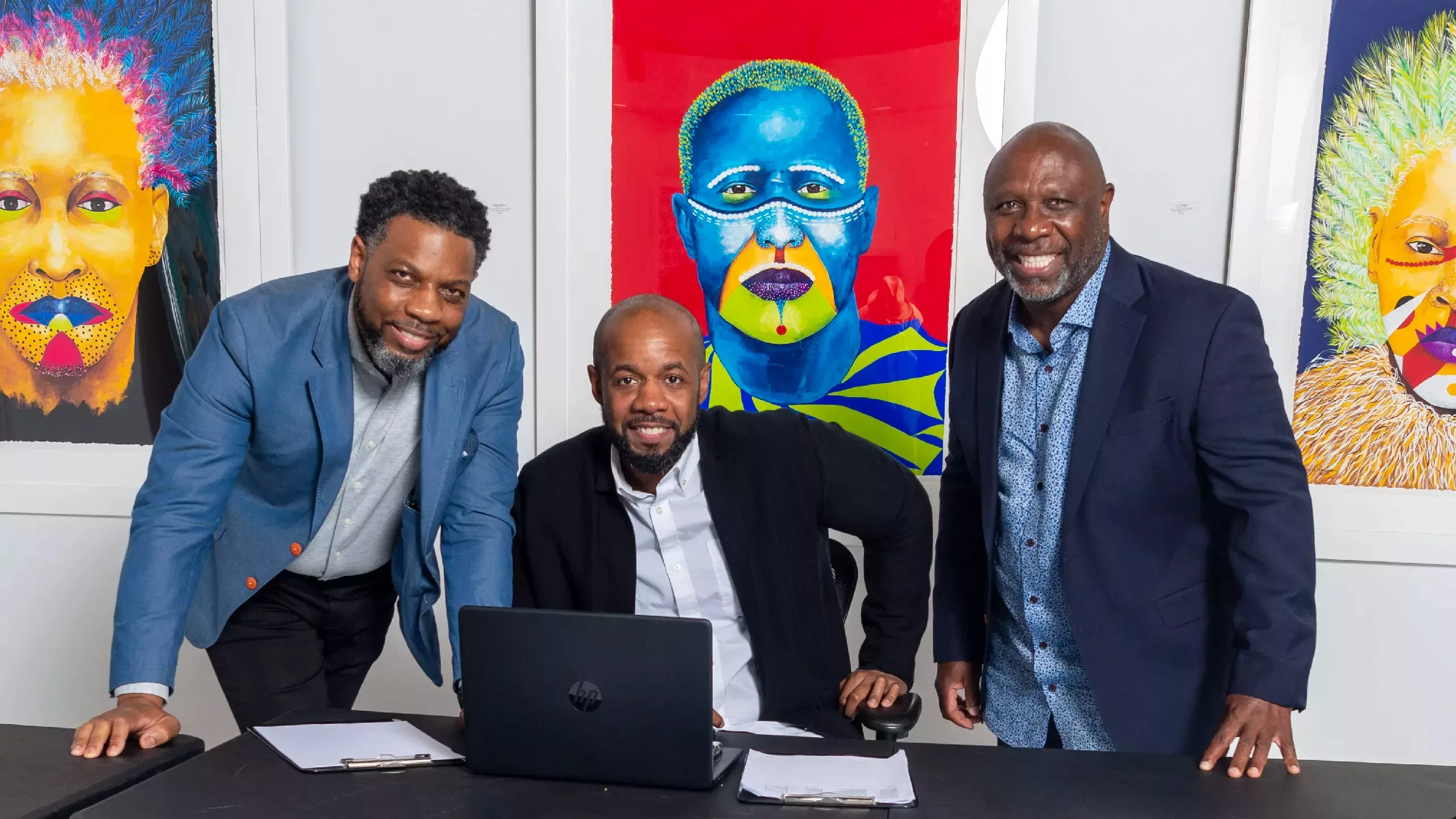 Three men gathered around a laptop smiling at the camera with art hanging in the background