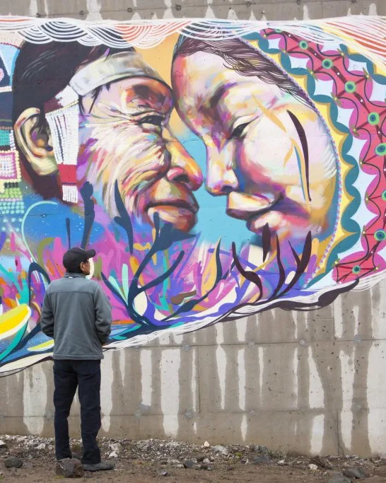 A man stands in front of a painted mural on a wall. The mural is a colorful depiction of two indigenous peoples from Iqaluit.