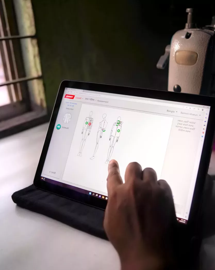 A Shimmy trainee touches her tablet screen during a digital tutorial. A sewing machine is seen in the background.