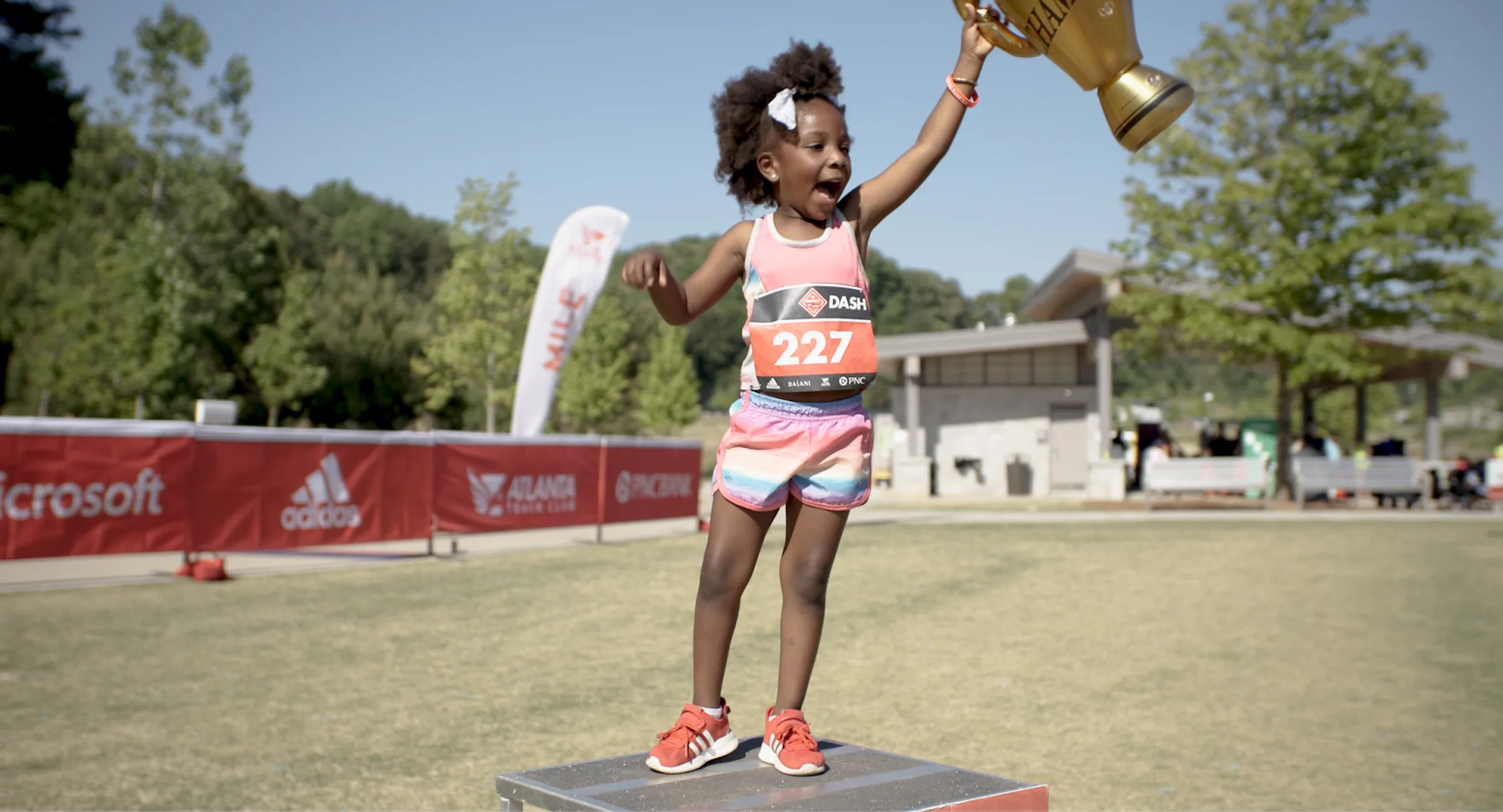 Young girl with running bib holding up a trophy on grass field.