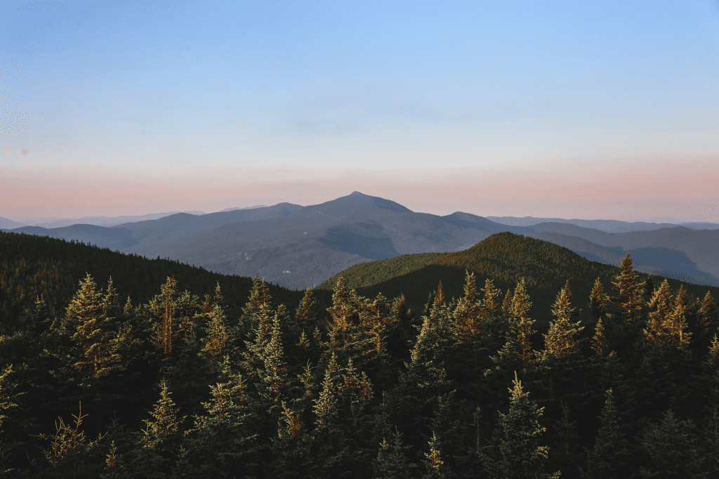 A view from above of a forest and rows of mountain peaks. The sky at the horizon is orange.