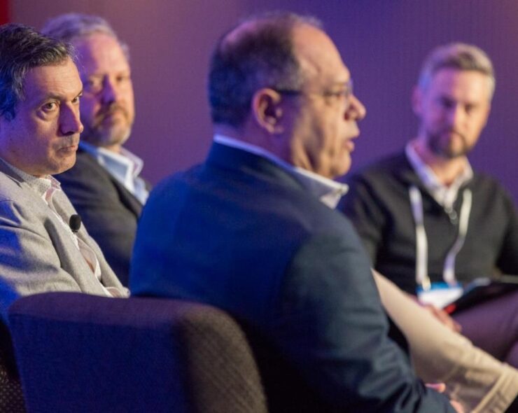 Four men sitting on stage, one is talking and the other three are looking at him listening