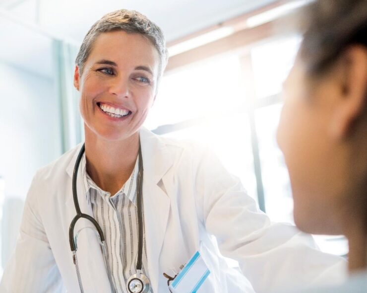 Smiling female doctor looking at patient. Focus is on confident practitioner wearing stethoscope. They are in hospital ward.