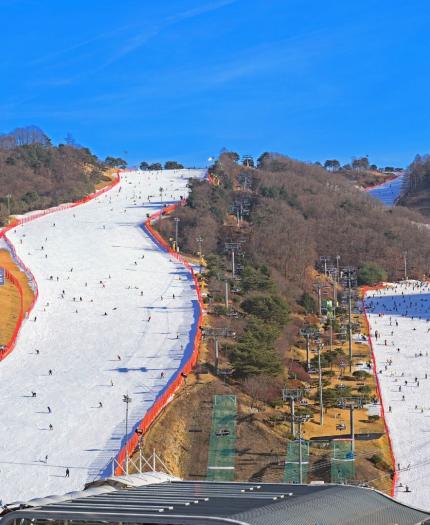 Aerial view of a snow-covered ski resort with multiple ski slopes surrounded by wooded hills, skiers and snowboarders visible on the slopes, and a clear blue sky above.