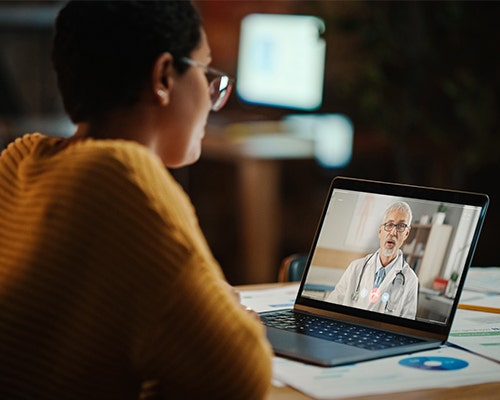 A person wearing glasses and sitting at a table is having a video call with a doctor displayed on a laptop screen.