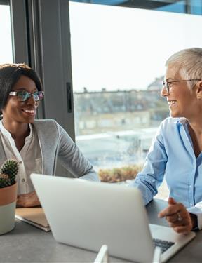 Two women sit at a table in an office setting holding a conversation with each other.