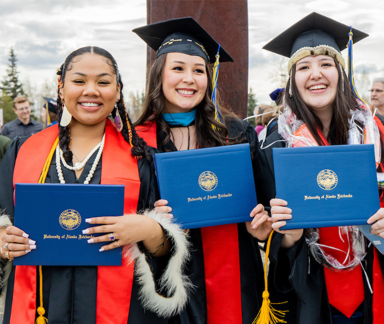 three UAF female graduates in regalia hold up their blue diplomas