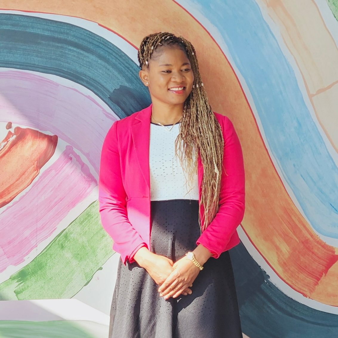 Dr. Rita Orji is wearing a pink cardigan and navy skirt, standing in front of a colourful mural.