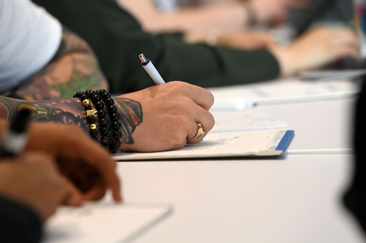 Close up of a student with a black bracelet and tattooed arm taking notes on a pad of paper.