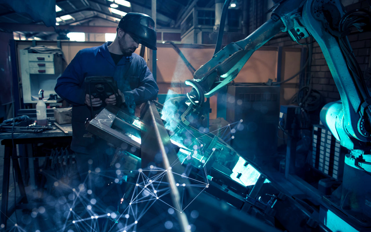 A worker with a welding helmet observes a robotic arm in a machine shop setting.