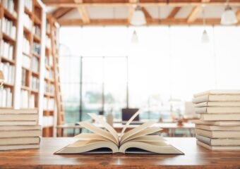 An open book on a table between two stacks of books, and library window in the background.