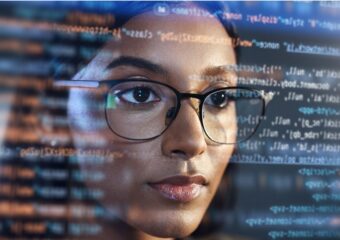 African-American woman wearing glasses reviews software coding. Image POV is from behind the coding looking out from inside the monitor.