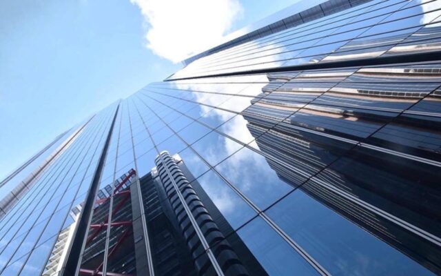 Reflections of high rise buildings appearing in the glass windows of another high rise building with a partly cloudy sky above.