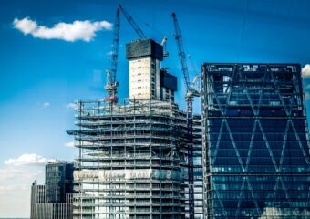 Building under construction with cranes in the background against a mostly sunny blue sky.