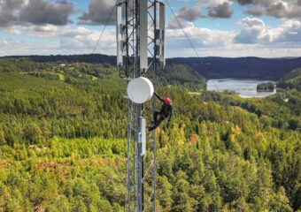 Field technician scales telecommunications tower with a forest and body of water in the background.