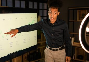 Young black man in business casual attire speaks to data on large screen for presentation.