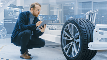 Man holding tablet inspects an automobile frame and wheel assembly in a factory.