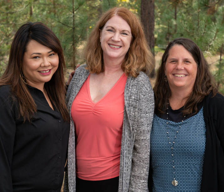 Three women pose for a photo in a forest area, they are standing close together and smiling