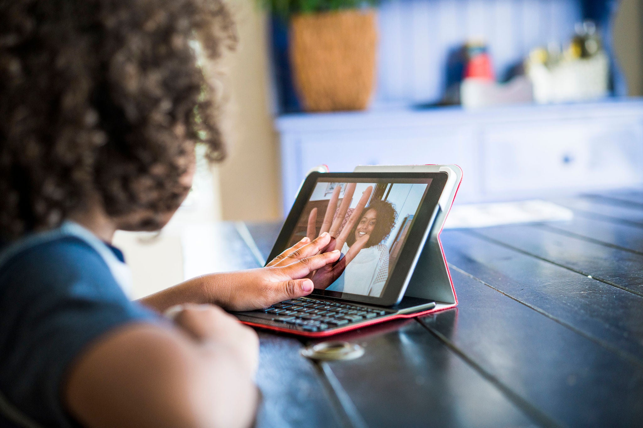 A photo of little girl video conferencing with mother on digital tablet. Cropped image of daughter is touching screen of tablet computer. She is at table in home.