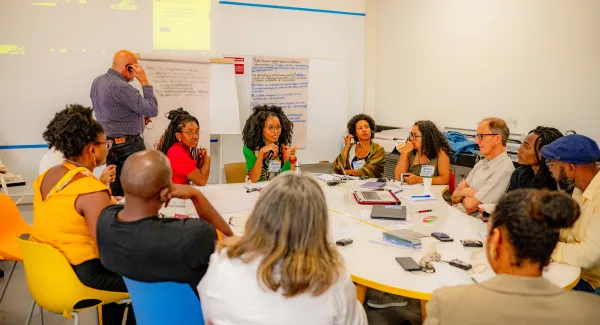 A diverse group of people sits around a large white table in a brightly lit room, engaged in a discussion. Some are talking while others listen attentively. A whiteboard in the background displays handwritten notes, and a laptop and papers are on the table.