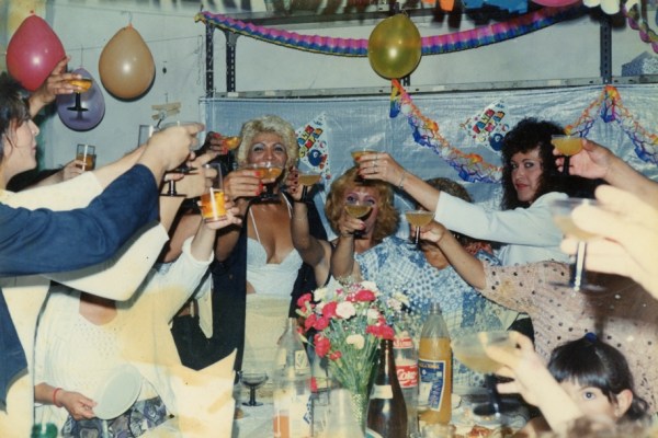 A flash photograph of people holding up glasses filled with orange drinks to cheers at a party. On the table are drinks, cups, and flowers. And there are balloons and streamers hanging on the back wall.