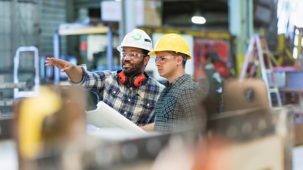 Two men wearing hard hats and safety gear stand in an industrial setting. One man is pointing while holding blueprints, and the other looks on attentively. The background includes various machinery and equipment.