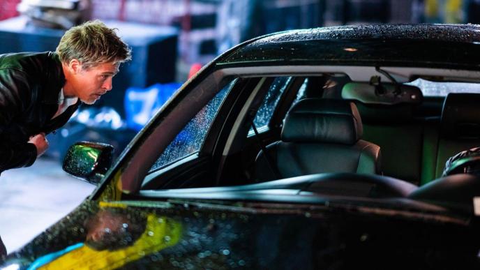 A man leaning down to talk to another man sat in a car, they are in a city in the rain.