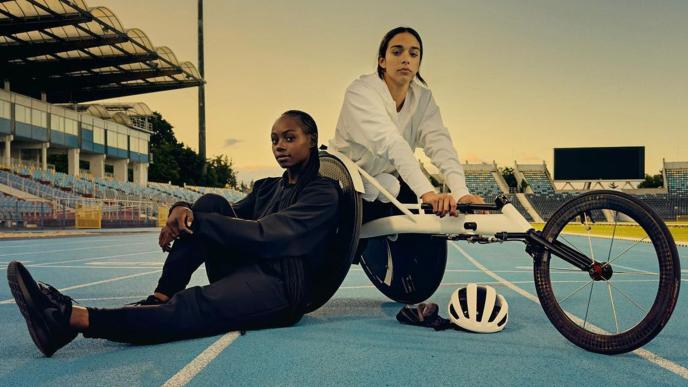 Two female athletes on a track; one of the athletes wears all black, one wears all white. The athlete in all white is in a wheelchair.  