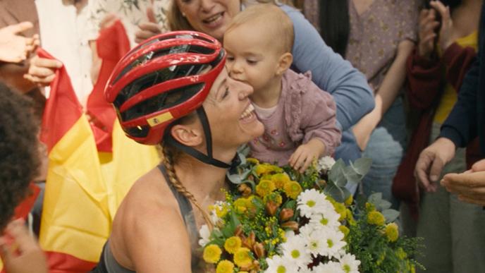 A Paralympian in her wheelchair bike holds a baby and a bouqet of flowers. A large group of loved ones and friends surround her after she completes her race. 