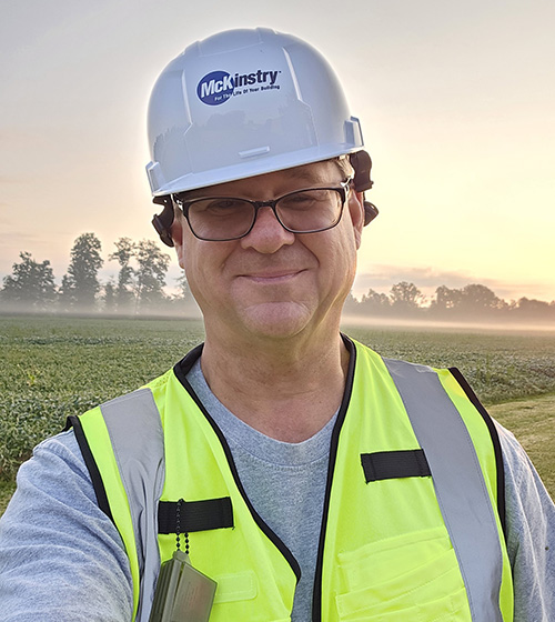 Photo of Kurt Holko McKinstry outdoors in front of a field wearing a hard hat and yellow safety vest.