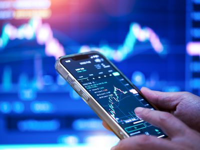 A close-up image of a person's hands holding a cell phone displaying stock market data, with a computer screen in the background.