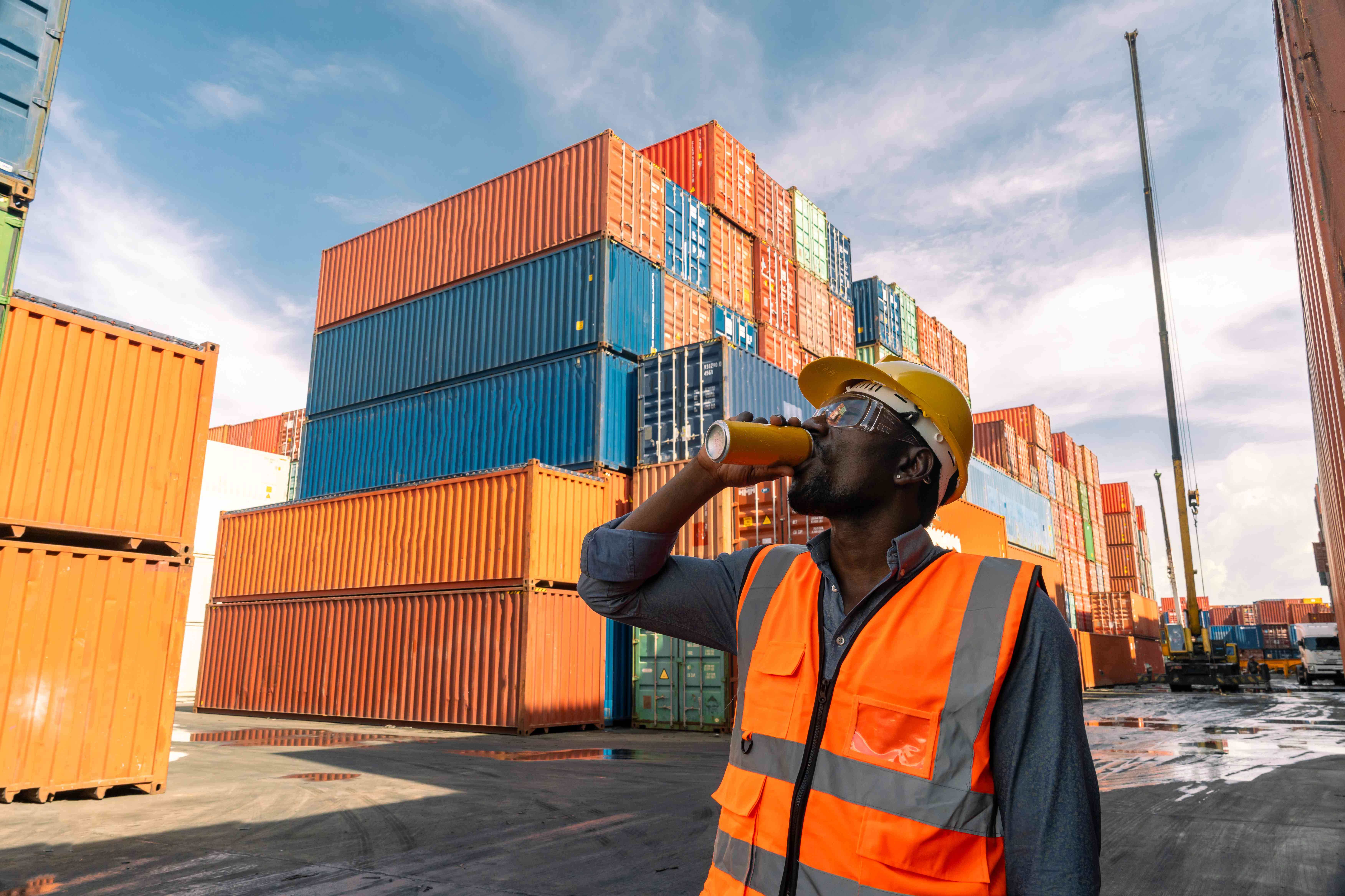 Worker stands amid shipping containers and drinks a canned drink while in the background a crane unloads more shipping containers.