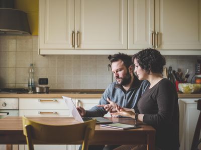 A man and a woman sit at a kitchen table looking at a laptop