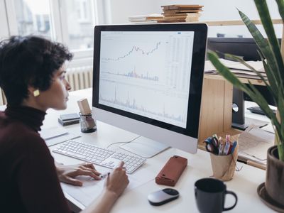 A woman sits at a desk, looking at a computer screen displaying stock figures and charts.