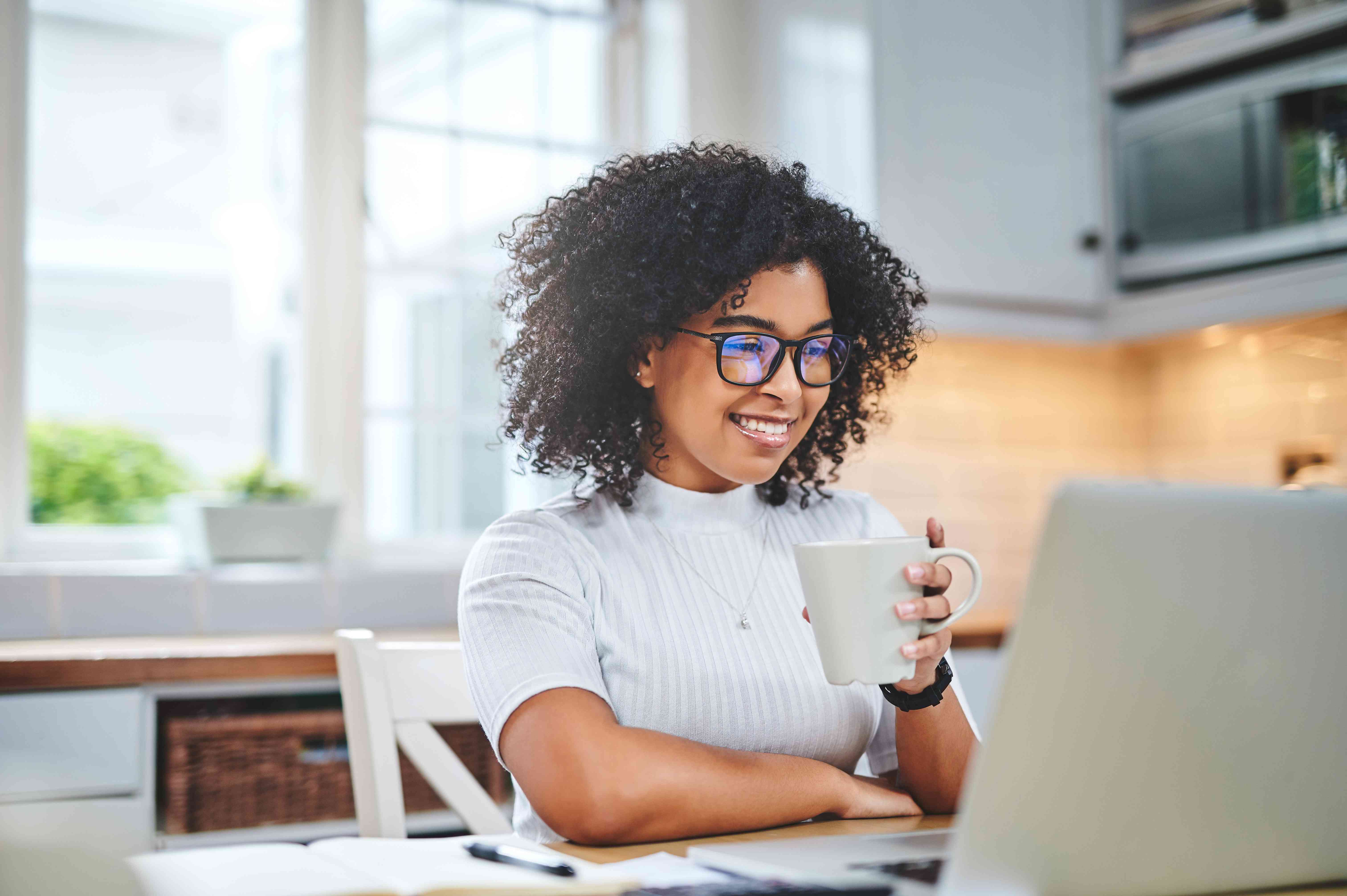 A young woman uses a laptop at her kitchen table, holding a coffee mug and smiling.